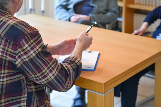 Jemand der an meinem Tisch sitzt mit Stift und Papier und dieser Person gegenüber sitzen auch 2 Menschen.